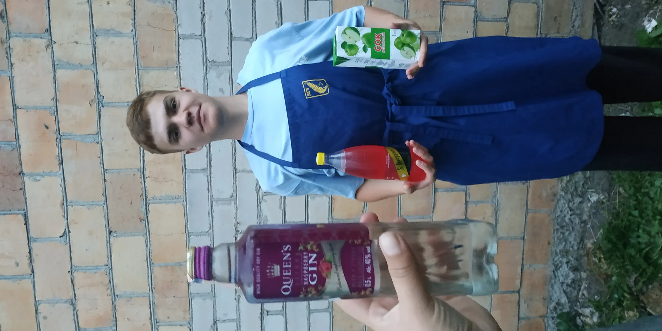 Парень в синем фартуке держит сок GOK и красную бутылку, на переднем плане бутылка Queen's Gin